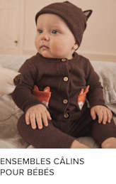 Un bébé souriant portant un bonnet et un pull en tricot marron, assis sur une surface douce avec un fond confortable et aux tons chauds.
