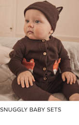 A smiling baby wearing a brown knitted hat and sweater, sitting on a soft surface with a cozy, warm-toned background.