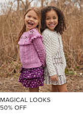 Two young girls, one with long blonde hair and the other with curly dark hair, are embracing and smiling joyfully in a field of tall grass.
