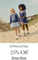 Two young girls, one with blonde hair and the other with curly dark hair, are walking together on a sandy beach with dunes in the background.