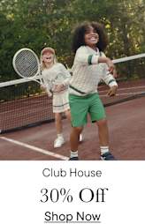 Two young children, a boy and a girl, are playing tennis on a tennis court surrounded by trees and greenery.