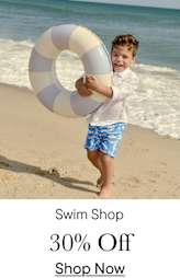 A young boy enthusiastically holds a white and blue striped life preserver on a sandy beach with the ocean visible in the background.