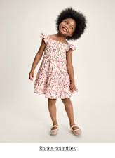 Une jeune fille aux cheveux noirs bouclés porte une robe rose à fleurs et sourit largement sur un fond uni.