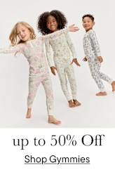 Tres niños pequeños, dos niñas y un niño, vestidos con pijamas estampados de colores, están de pie juntos y sonriendo sobre un fondo blanco brillante.