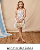 Une jeune fille aux cheveux blonds bouclés se tient debout sur un parquet, vêtue d'une robe à motifs et tenant un sac à main en paille, sur un fond beige.