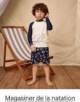 Un jeune enfant aux cheveux bouclés se tient pieds nus sur un plancher en bois, vêtu d'une chemise blanche et bleu marine et d'un short à motifs bleu marine, à côté d'une chaise en bois.