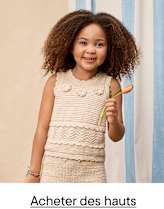 Une jeune fille aux cheveux bruns bouclés et au sourire chaleureux, vêtue d'une robe beige en crochet, se tient devant un fond bleu pastel et blanc.