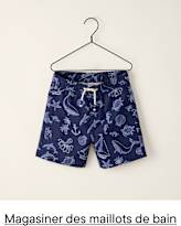 Un cintre en fil de fer noir, muni d'une extrémité en forme de cœur, maintient un maillot de bain rayé à petits motifs en forme de cœur sur fond blanc uni.