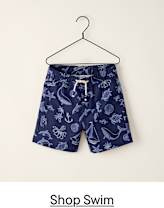 A navy blue pair of swim trunks with a white drawstring and a pattern of sea creatures and nautical elements hangs on a black hanger against a plain, light-colored background.