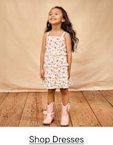 A young girl in a floral dress and pink boots stands on a wooden floor against a beige backdrop.