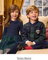 Two smiling children sit on a beige sofa in a room with a window and gold curtains.