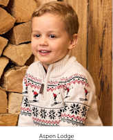 A young boy wearing a white patterned sweater stands in front of a stack of logs and a wooden wall.