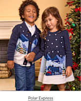 Two children wearing navy blue Polar Express themed outfits stand together in front of a fireplace and a decorated Christmas tree.
