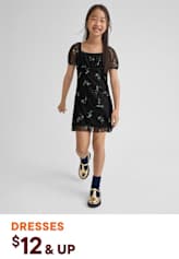 A young girl wearing a black floral dress and patterned shoes stands against a plain background, smiling brightly.