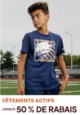Un jeune portant un t-shirt bleu marine avec un motif graphique se tient sur un terrain de sport avec une piste et des poteaux de but visibles en arrière-plan.