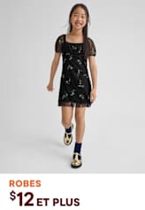 Une jeune fille portant une robe noire à fleurs et des chaussures à motifs se tient debout sur un fond uni, souriant vivement.