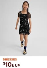A young girl wearing a black floral dress and patterned shoes stands against a plain background, smiling brightly.