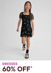 A young girl wearing a black floral dress and patterned shoes stands against a plain background, smiling brightly.