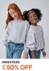 Two young girls, one with red hair and the other with curly black hair, are standing together and smiling. They are wearing cozy, patterned sweaters and jeans, and the background is a plain white studio setting.