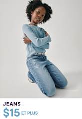 Une jeune fille portant une robe noire à fleurs et des chaussures à motifs se tient debout sur un fond uni, souriant vivement.