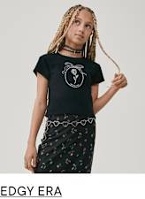 A young person with braided hair wearing a black t-shirt with a graphic design and a floral patterned skirt, standing against a plain background.