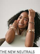 A smiling young woman with curly dark hair and jewelry adorning her arms sits against a plain background.