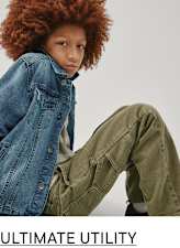 The image features a person with curly red hair wearing a denim jacket and olive green pants, sitting against a plain white background.