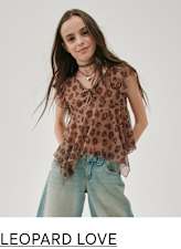 Una mujer joven con cabello largo y castaño, que viste una blusa con estampado de leopardo y jeans de color claro, está parada frente a un fondo liso.