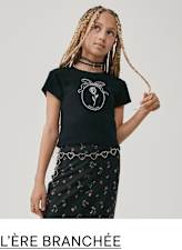 Un jeune aux cheveux tressés portant un t-shirt noir avec un motif graphique et une jupe à motifs floraux, debout sur un fond uni.