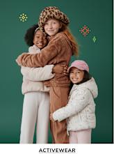 Three young girls wearing warm, cozy outfits are embracing each other in front of a solid green background, creating a cheerful and affectionate scene.