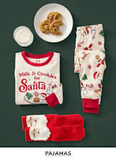 A festive arrangement featuring a plate of cookies, a milk glass, a "Milk & Cookies for Santa" t-shirt, and a pair of Christmas-themed pajamas on a dark green background.