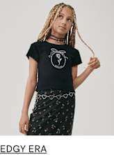 A young person with braided hair wearing a black t-shirt with a graphic design and a floral patterned skirt, standing against a plain background.