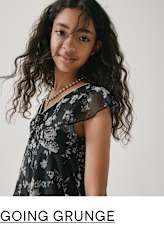 A young woman with curly dark hair wearing a floral print dress stands against a plain white background, her expression conveying a casual, grunge-inspired style.