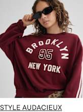 Une jeune femme portant un sweat-shirt bordeaux avec l'inscription « Brooklyn 95 New York » imprimée dessus, associé à un pantalon à imprimé léopard, se tient debout sur un fond uni tout en portant des lunettes de soleil.