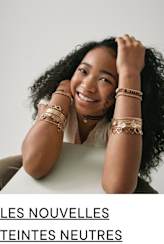 Une jeune femme souriante aux cheveux noirs bouclés et aux bijoux ornant ses bras est assise sur un fond uni.