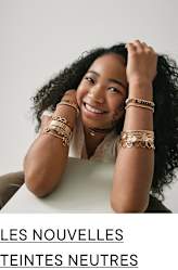 Une jeune femme souriante aux cheveux noirs bouclés et aux bijoux ornant ses bras est assise sur un fond uni.