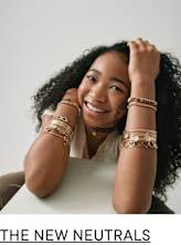 A smiling young woman with curly dark hair and jewelry adorning her arms sits against a plain background.
