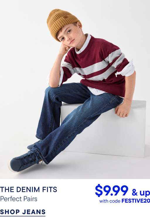 A young boy wearing a mustard beanie, a red and grey striped sweater, and blue jeans sits on a white block against a plain white background.