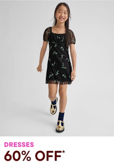 A young girl wearing a black floral dress and navy blue and white patterned shoes stands against a plain gray background, smiling brightly.