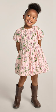 Una niña pequeña con un vestido rosa estampado de flores y botas marrones posa sobre un fondo liso de color claro.