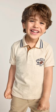 Un niño pequeño sonriente, vestido con una camisa polo color crema y pantalones color canela, posa sobre un fondo liso y claro.