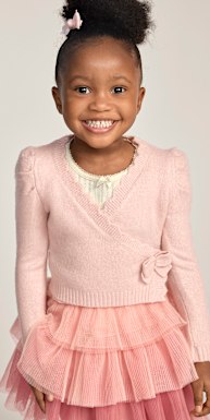 Una niña sonriente, vestida con un cárdigan rosa y un tutú, posa sobre un fondo liso y claro.