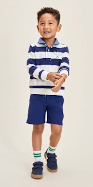 Un niño pequeño que viste una camiseta polo a rayas y pantalones cortos azul marino se encuentra frente a un fondo sencillo y sonríe con confianza.