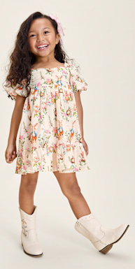 Una joven de cabello rizado lleva un vestido con estampado floral y botas de vaquero blancas, está de pie sobre un fondo beige liso y sonríe alegremente.