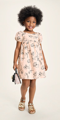 Una joven de cabello oscuro y rizado lleva un vestido con estampado floral y sandalias, de pie frente a un fondo blanco liso.