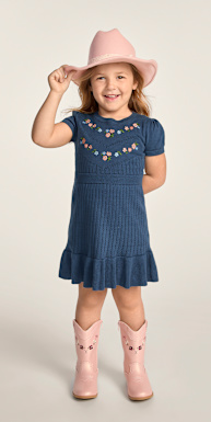 A smiling young girl in a blue dress and pink cowboy boots stands against a plain, light background, holding the brim of her pink hat.