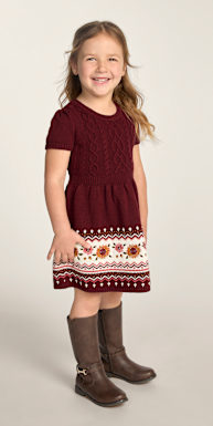 A smiling young girl in a maroon cable-knit dress and brown boots stands against a plain, light gray background.