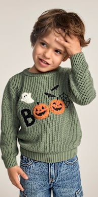 A young boy in a green Halloween-themed sweater and denim shorts stands against a plain, light gray background.