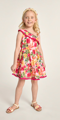 Una jovencita con cabello rubio largo y rizado lleva un colorido vestido floral y sandalias, posa frente a un fondo blanco liso y sonríe radiantemente.