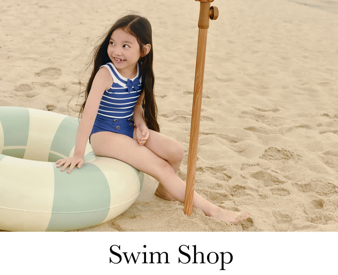 A young girl with long, dark hair is smiling joyfully on a sandy beach, with the ocean visible in the background.